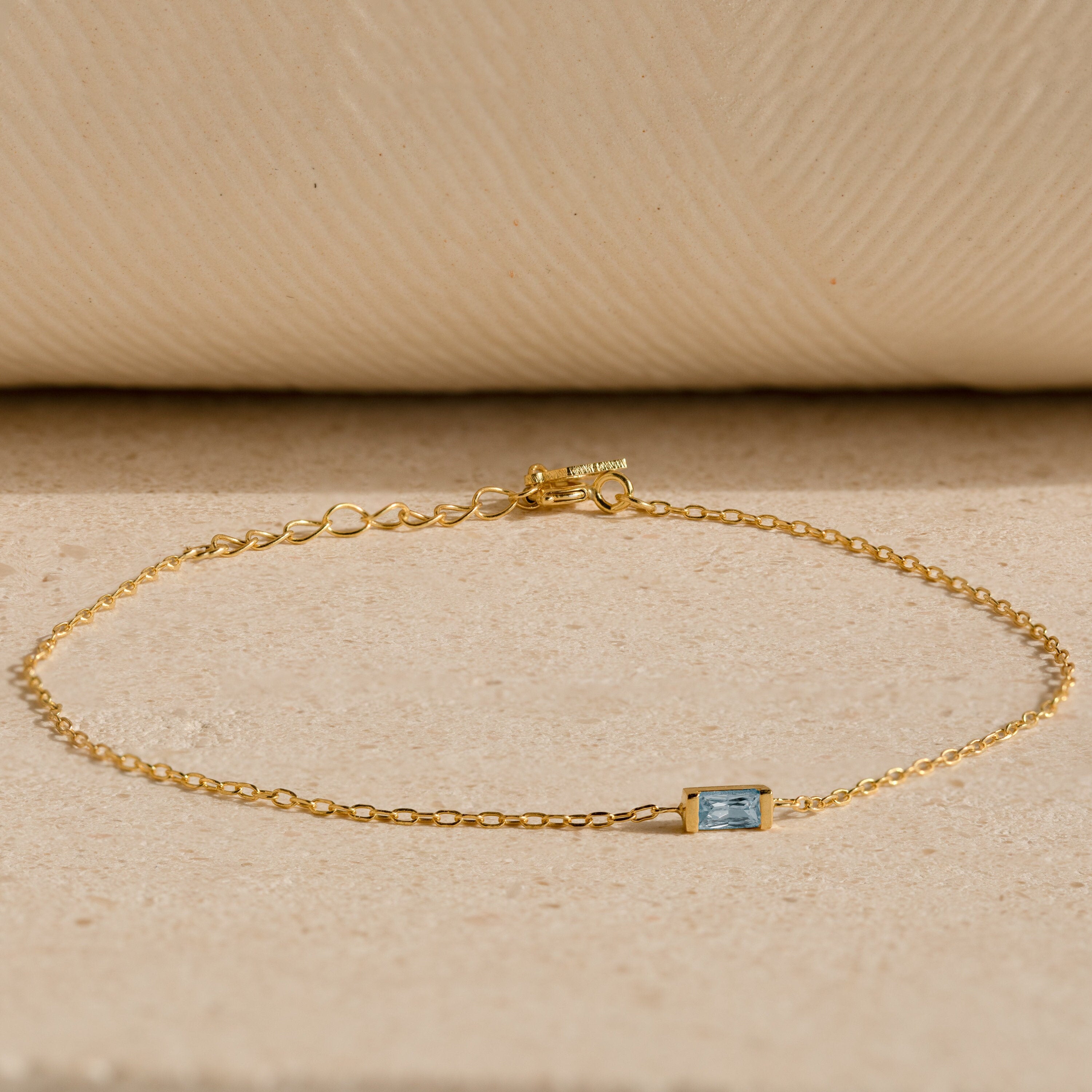 The Baguette Birthstone Bracelet features a delicate gold chain and a small rectangular blue gemstone, shown on a beige textured surface.