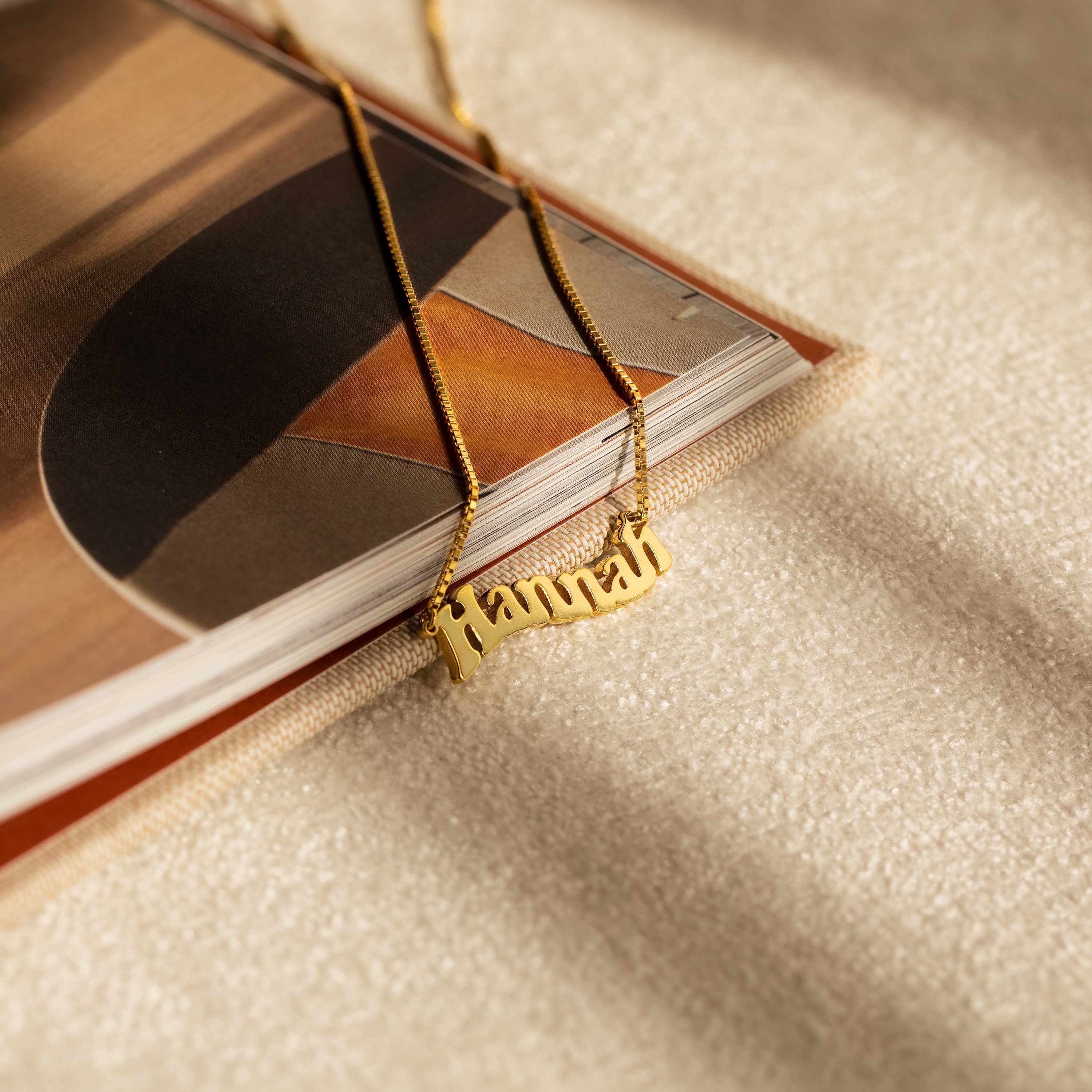 The Retro Name Necklace in Box Chain featuring "Hannah" rests on an open book under gentle sunlight, highlighting the classic appeal of personalized jewelry.