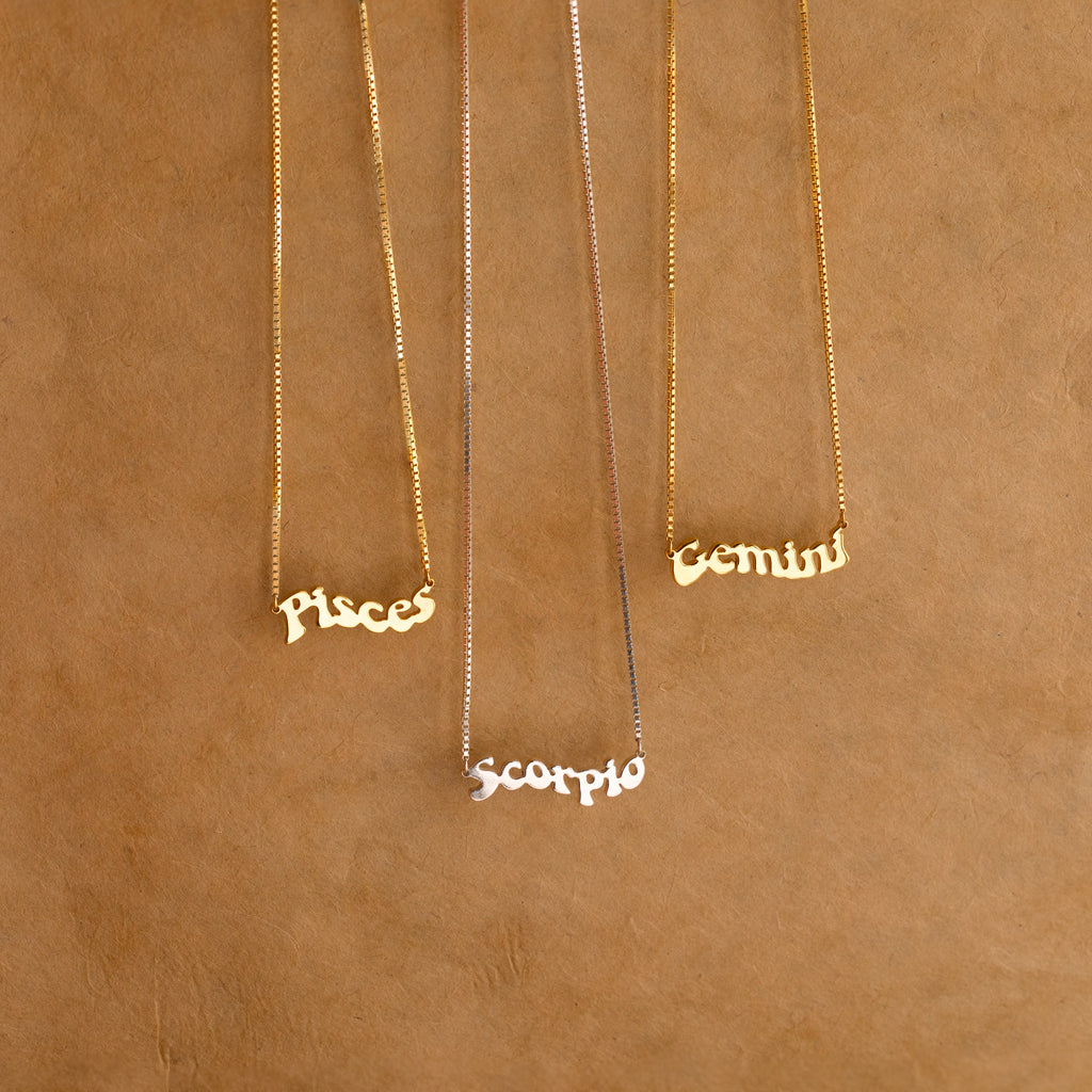 Three Retro Name Necklaces in Box Chain with Pisces, Scorpio, and Gemini pendants rest on a brown textured background—ideal custom charm jewelry for anyone who loves personalized pieces.
