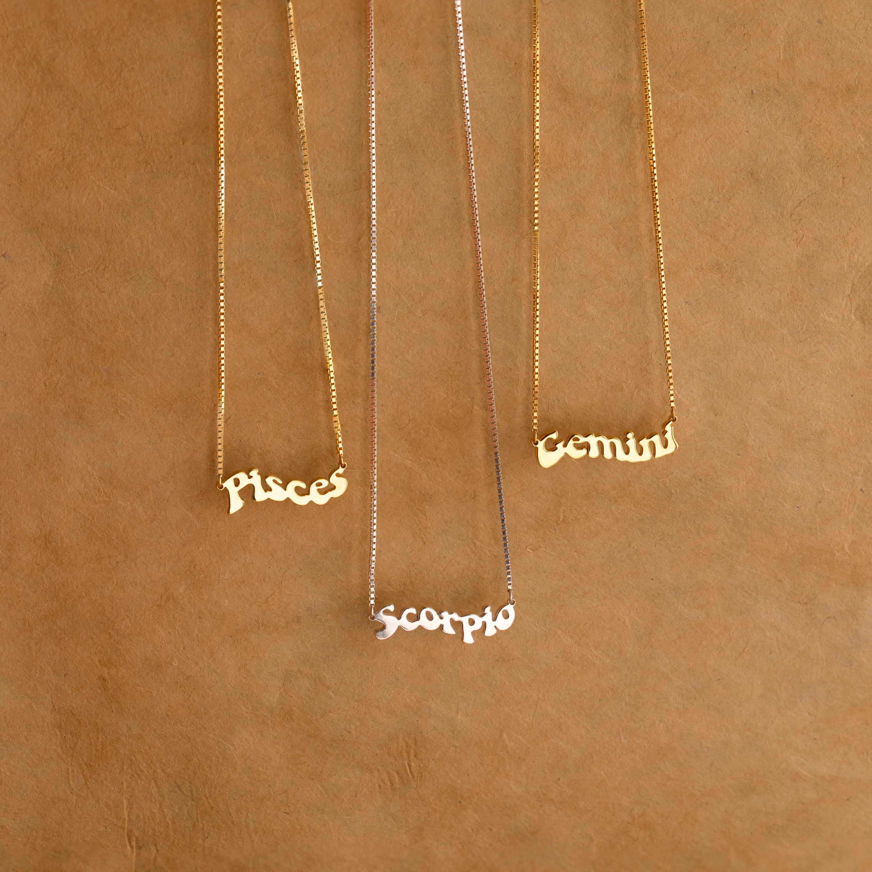 Three Retro Name Necklaces in Box Chain with Pisces, Scorpio, and Gemini pendants rest on a brown textured background—ideal custom charm jewelry for anyone who loves personalized pieces.