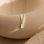 The Tiny Initial Tag Necklace features a gold chain with a rectangular RNS-engraved pendant, elegantly displayed on a cream surface—a classic piece of personalized jewelry for those seeking an initial necklace.