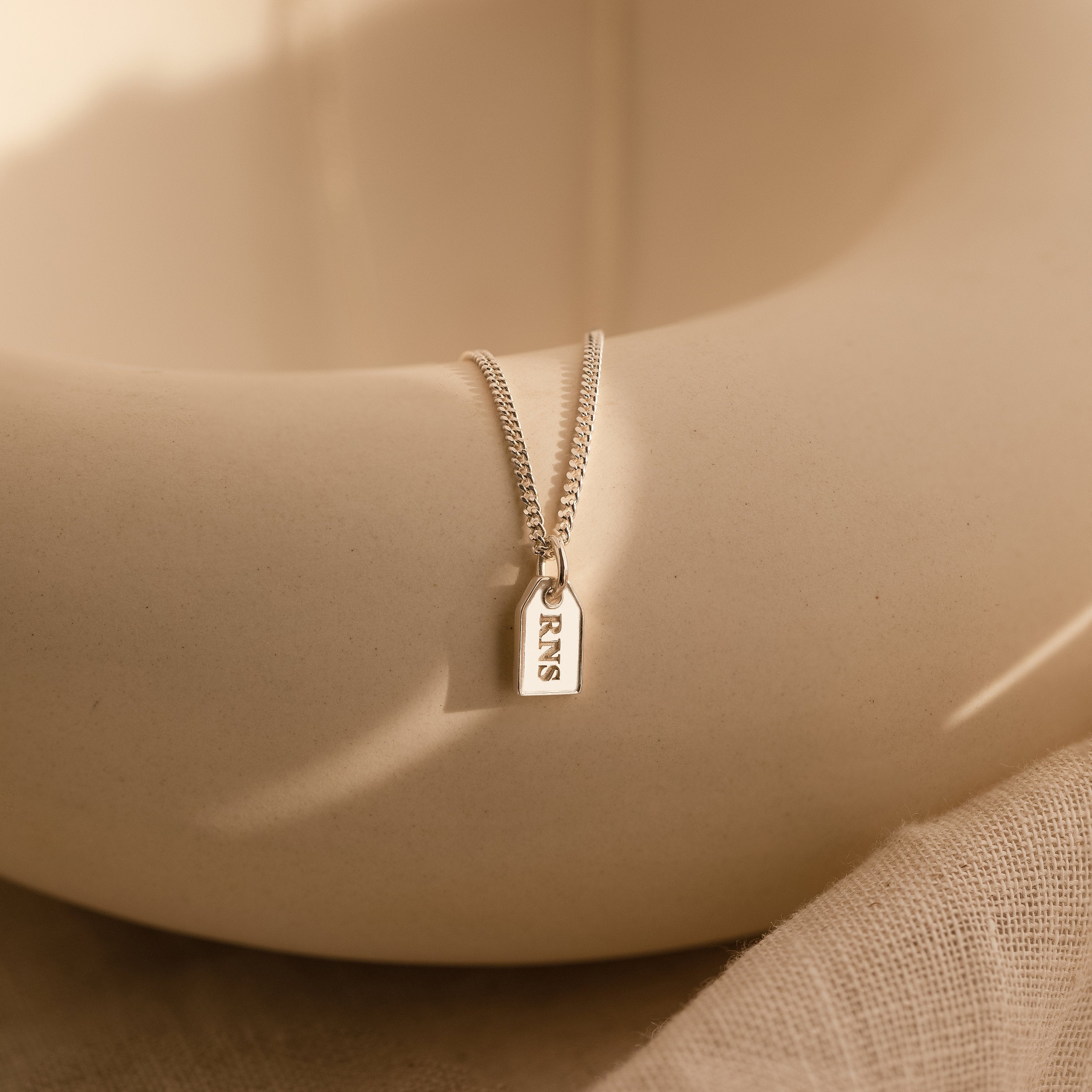 The Tiny Initial Tag Necklace features a gold chain with a rectangular RNS-engraved pendant, elegantly displayed on a cream surface—a classic piece of personalized jewelry for those seeking an initial necklace.