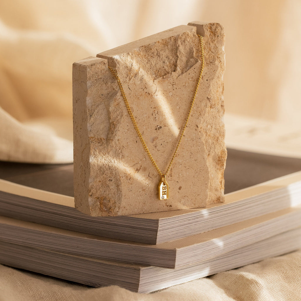 The Tiny Initial Tag Necklace features a sleek gold rectangular pendant, displayed elegantly on a textured stone atop stacked books—a timeless piece of personalized jewelry.