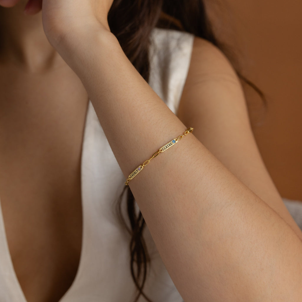 Woman wearing a Mini Name Birthstone Bracelet with an engraved bar, paired with a white sleeveless top against a brown background—a perfect personalized gift.