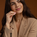 A woman in a beige blazer smiles gently, holding the Initial Heart Necklace—a custom piece featuring a circular pendant—against a brown background, making it an ideal gift for someone you love.