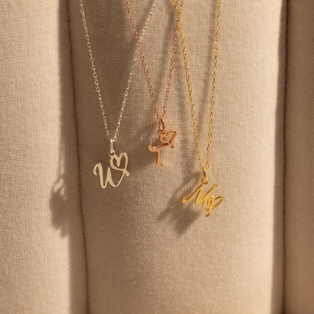 Three Initial Heart Necklaces in gold and silver, each with W, T, and M letter pendants, hang against beige fabric—a thoughtful gift for someone special.