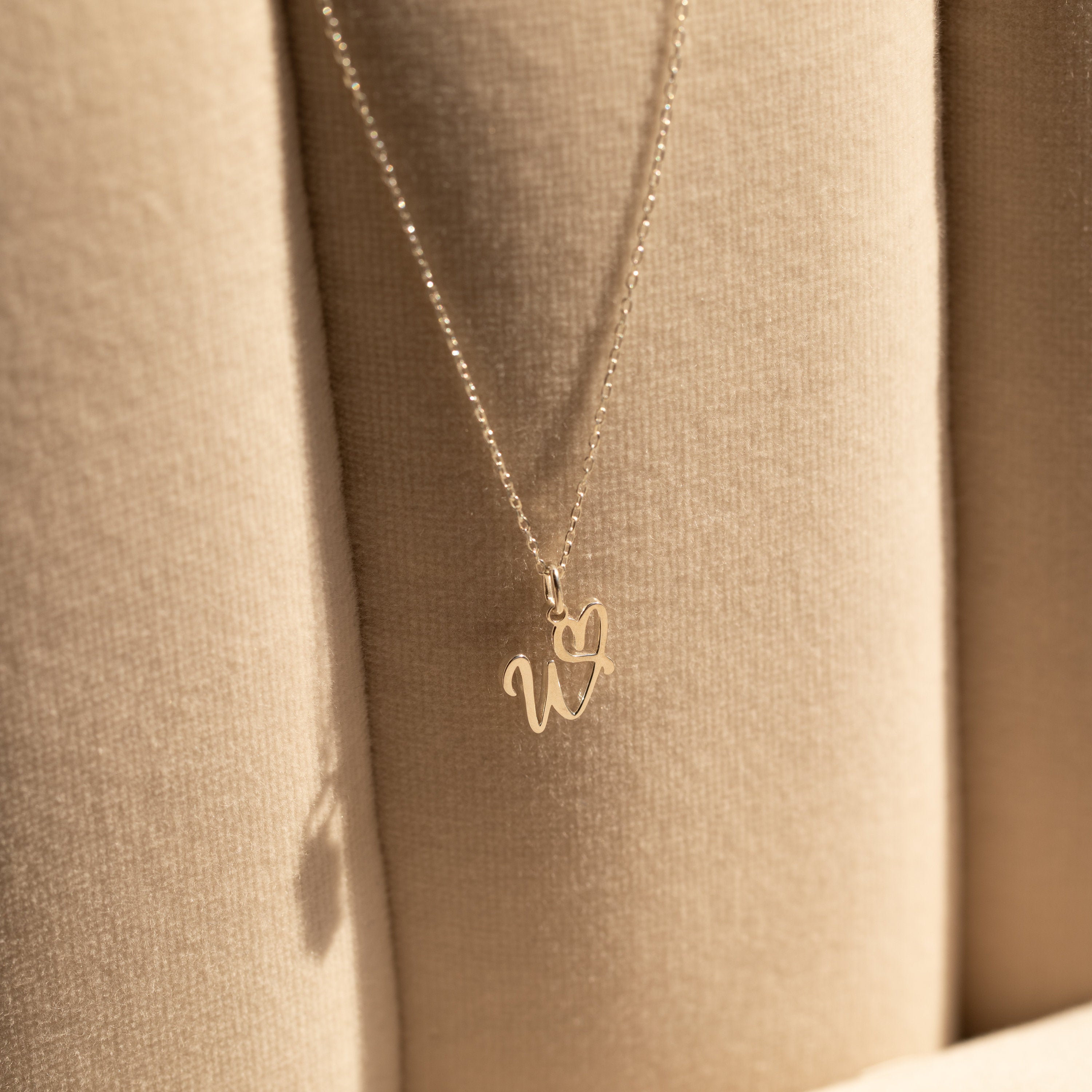 The Initial Heart Necklace, featuring a gold "w" and heart pendant, rests on a beige cushioned surface—an ideal gift to show someone you care.