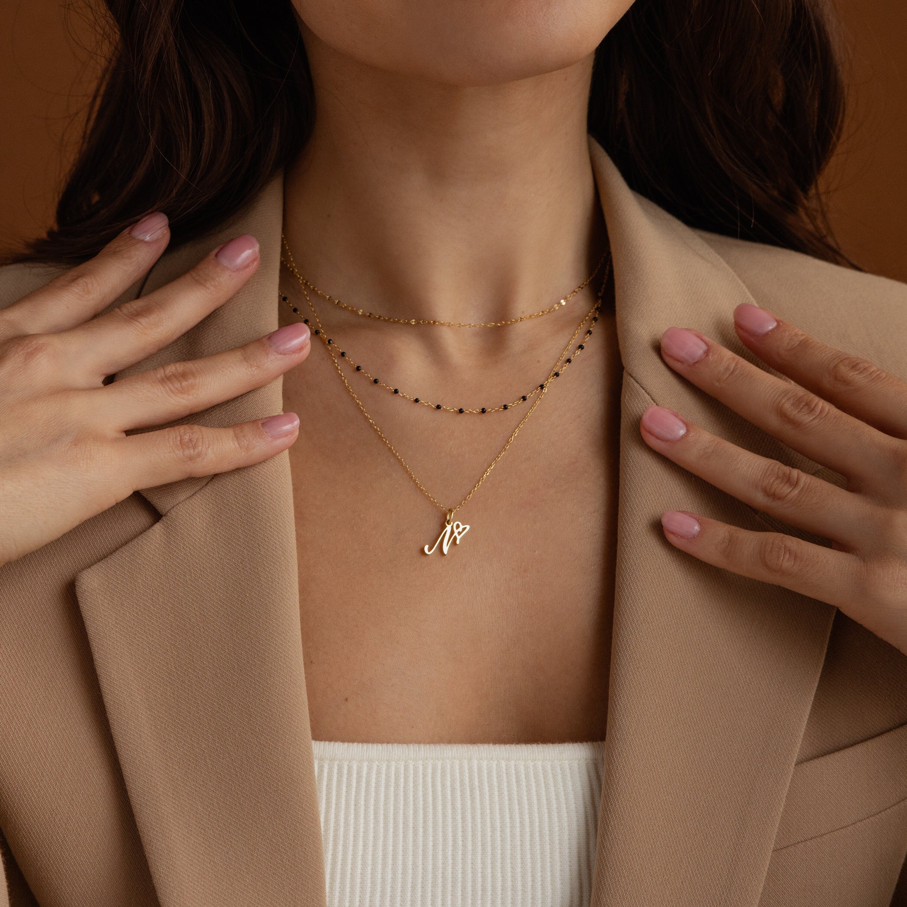 A woman wearing an Initial Heart Necklace and layered gold necklaces with a tan blazer, showcasing manicured nails—the Initial Heart Necklace is the perfect gift for someone you love.