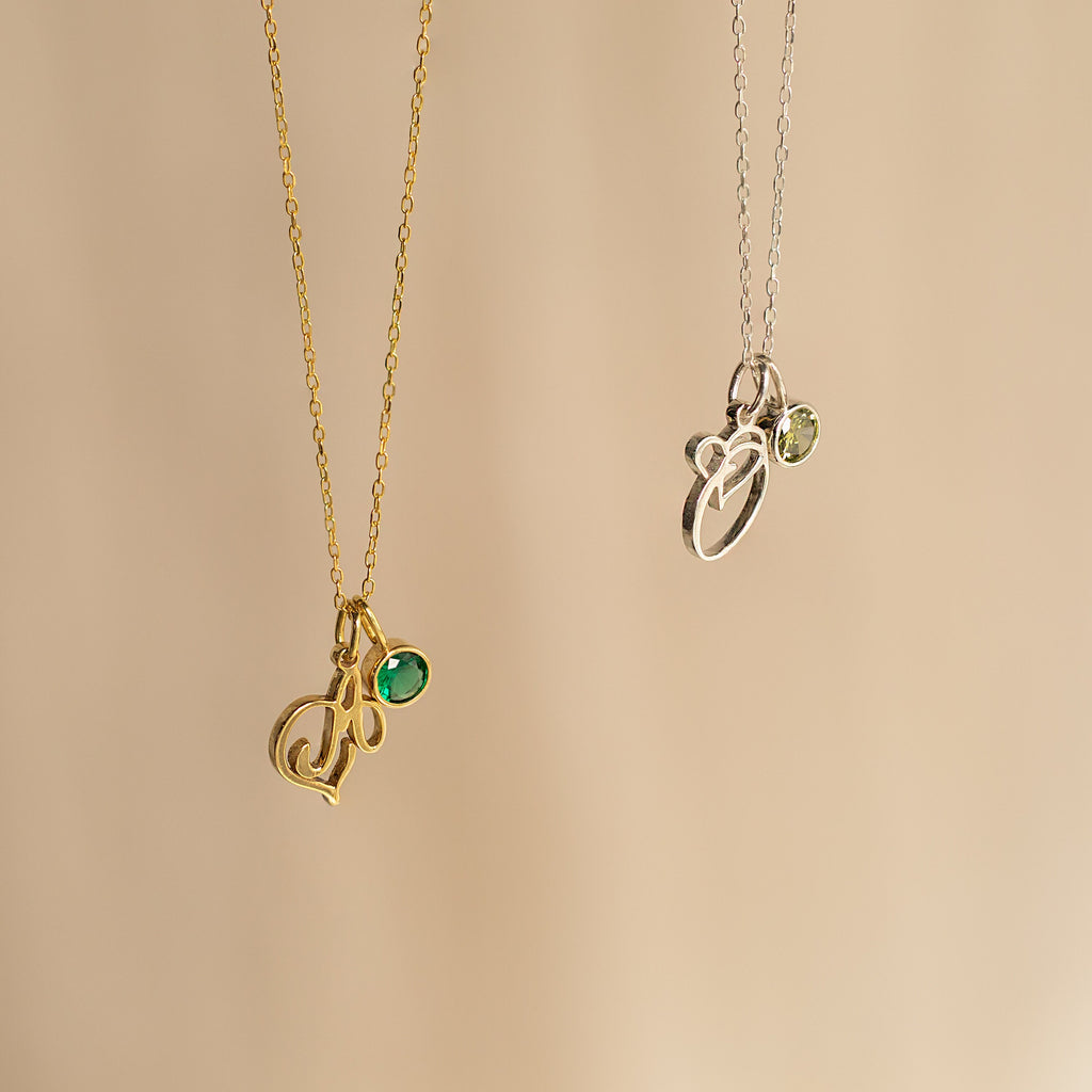 Two Initial Heart Birthstone Necklaces—one gold with a green gem, one silver with a light green gem—displayed on beige. A perfect personalized or custom birthstone gift to celebrate someone special.