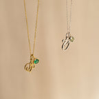 Two Initial Heart Birthstone Necklaces—one gold with a green gem, one silver with a light green gem—displayed on beige. A perfect personalized or custom birthstone gift to celebrate someone special.