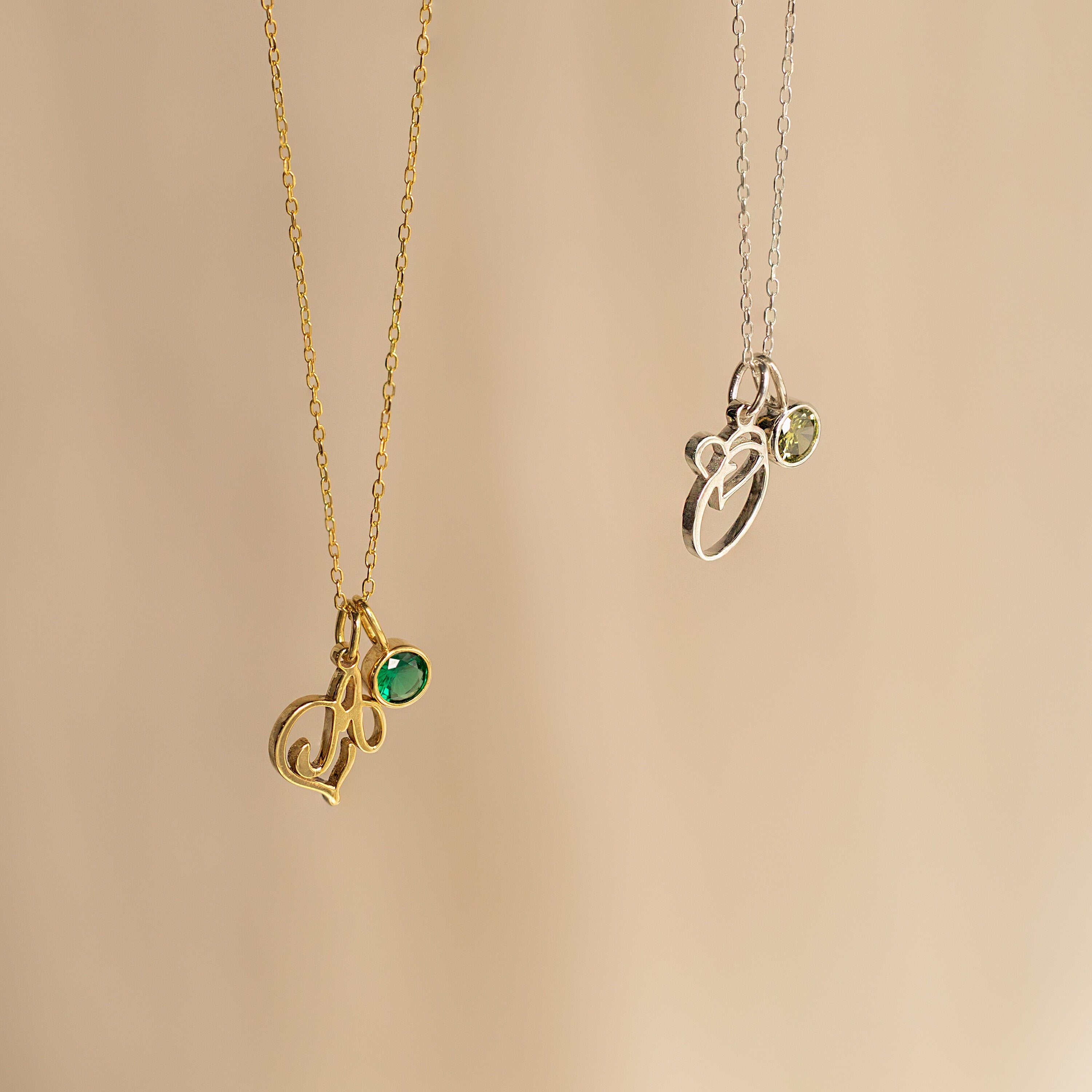 Two Initial Heart Birthstone Necklaces—one gold with a green gem, one silver with a light green gem—displayed on beige. A perfect personalized or custom birthstone gift to celebrate someone special.
