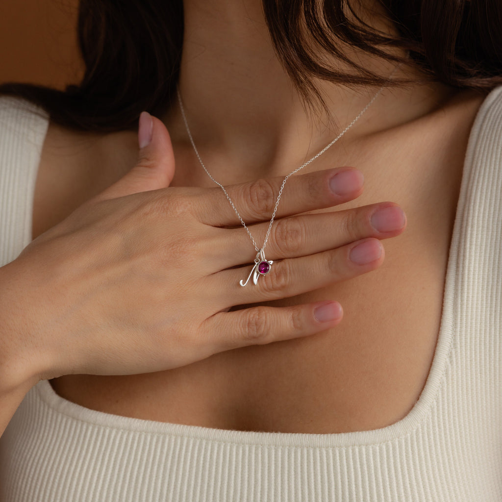 A woman touches her chest, wearing the Initial Heart Birthstone Necklace with a custom 'M' pendant and a small red birthstone.