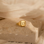The Lucky Number Signet Ring in gold, featuring the number 13, is displayed on a textured beige stone with soft fabric behind it—a bold statement piece for anyone seeking unique style.