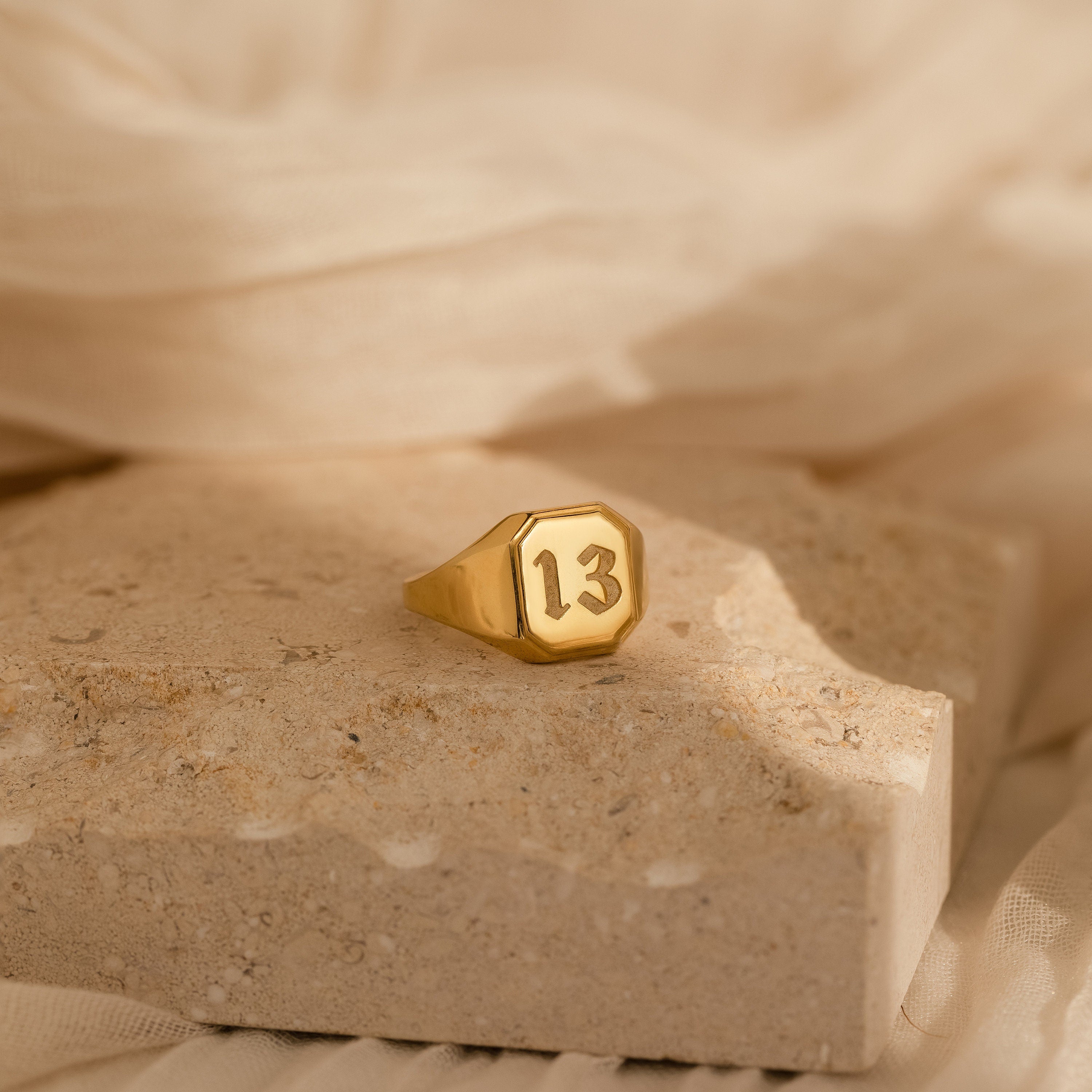The Lucky Number Signet Ring in gold, featuring the number 13, is displayed on a textured beige stone with soft fabric behind it—a bold statement piece for anyone seeking unique style.