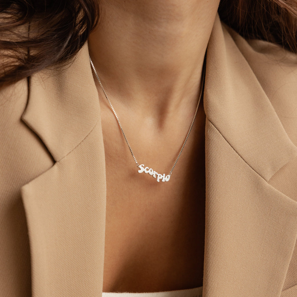 A woman wearing a beige blazer and a Retro Name Necklace in Box Chain with a silver Scorpio pendant.