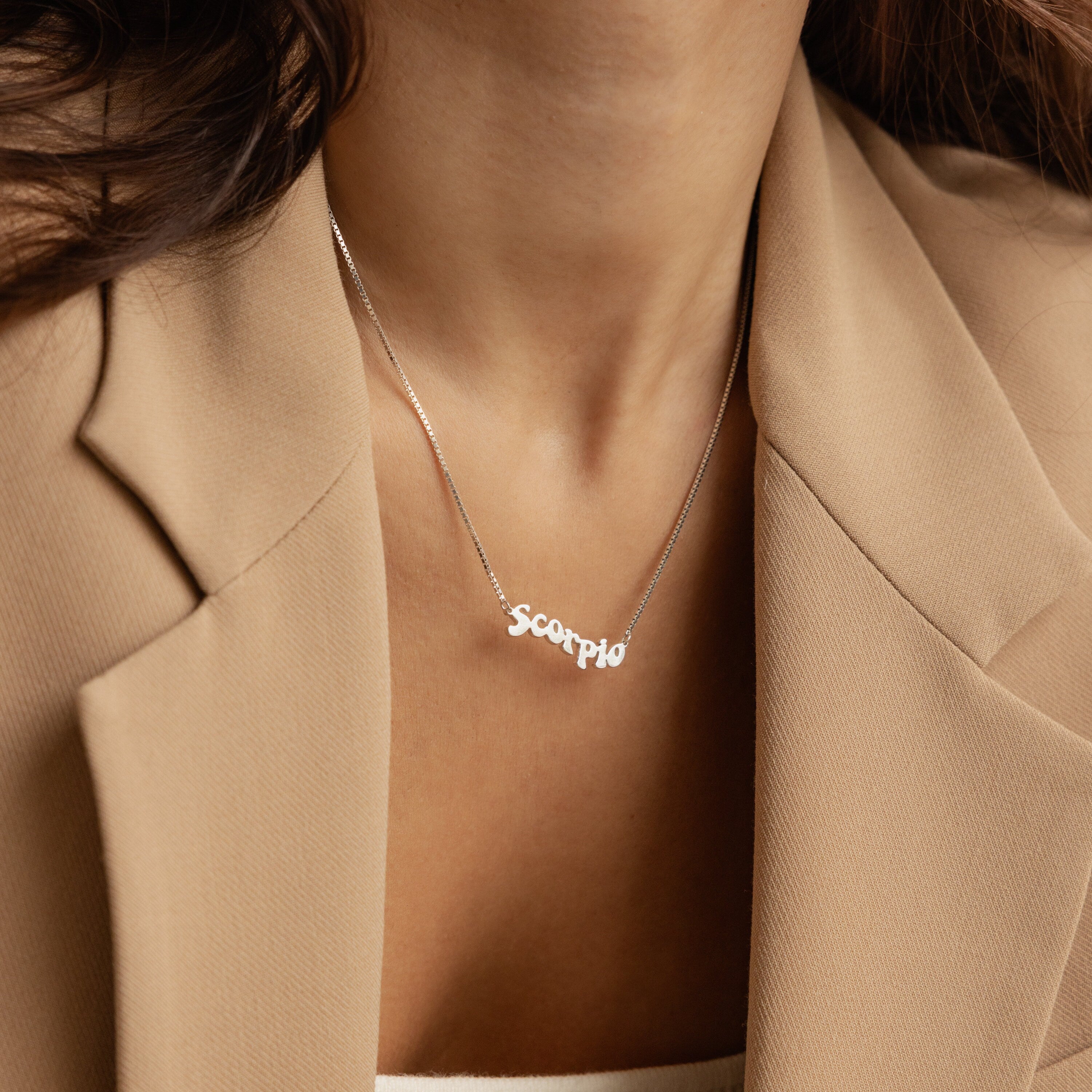 A woman wearing a beige blazer and a Retro Name Necklace in Box Chain with a silver Scorpio pendant.