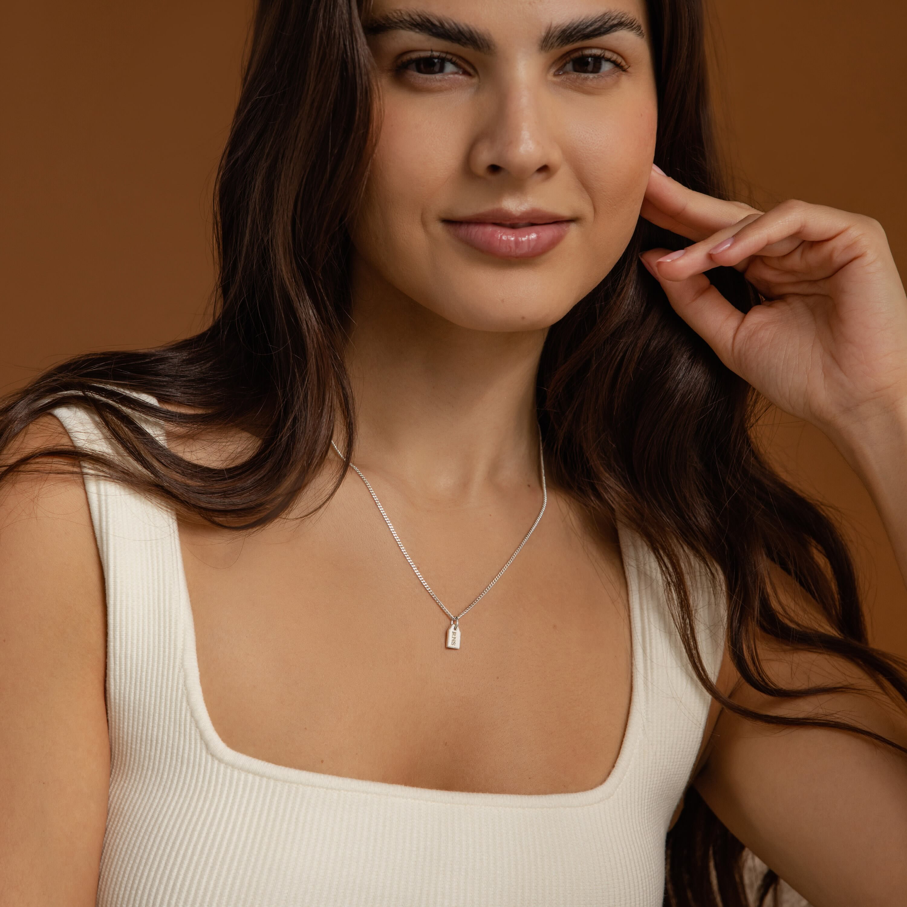 A woman with long brown hair, dressed in a square-neck ivory top, wears the Tiny Initial Tag Necklace and smiles slightly.