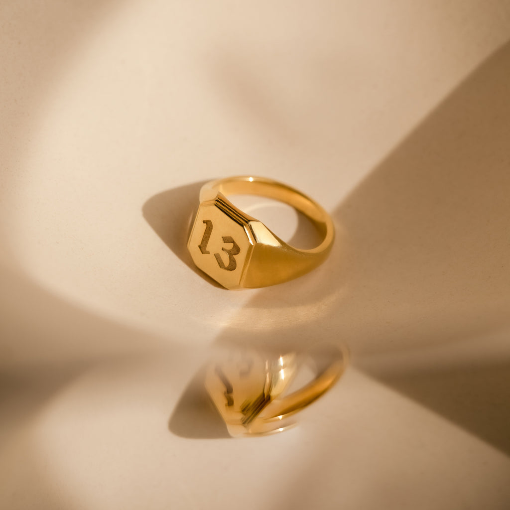 The Lucky Number Signet Ring features the number 13 engraved, displayed on a beige surface with soft shadows—ideal statement jewelry for fans of personalized engraving.
