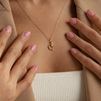 A woman wears the Initial Heart Necklace—an ideal gift for a loved one—styled with a gold heart pendant, beige blazer, and light pink manicured nails.