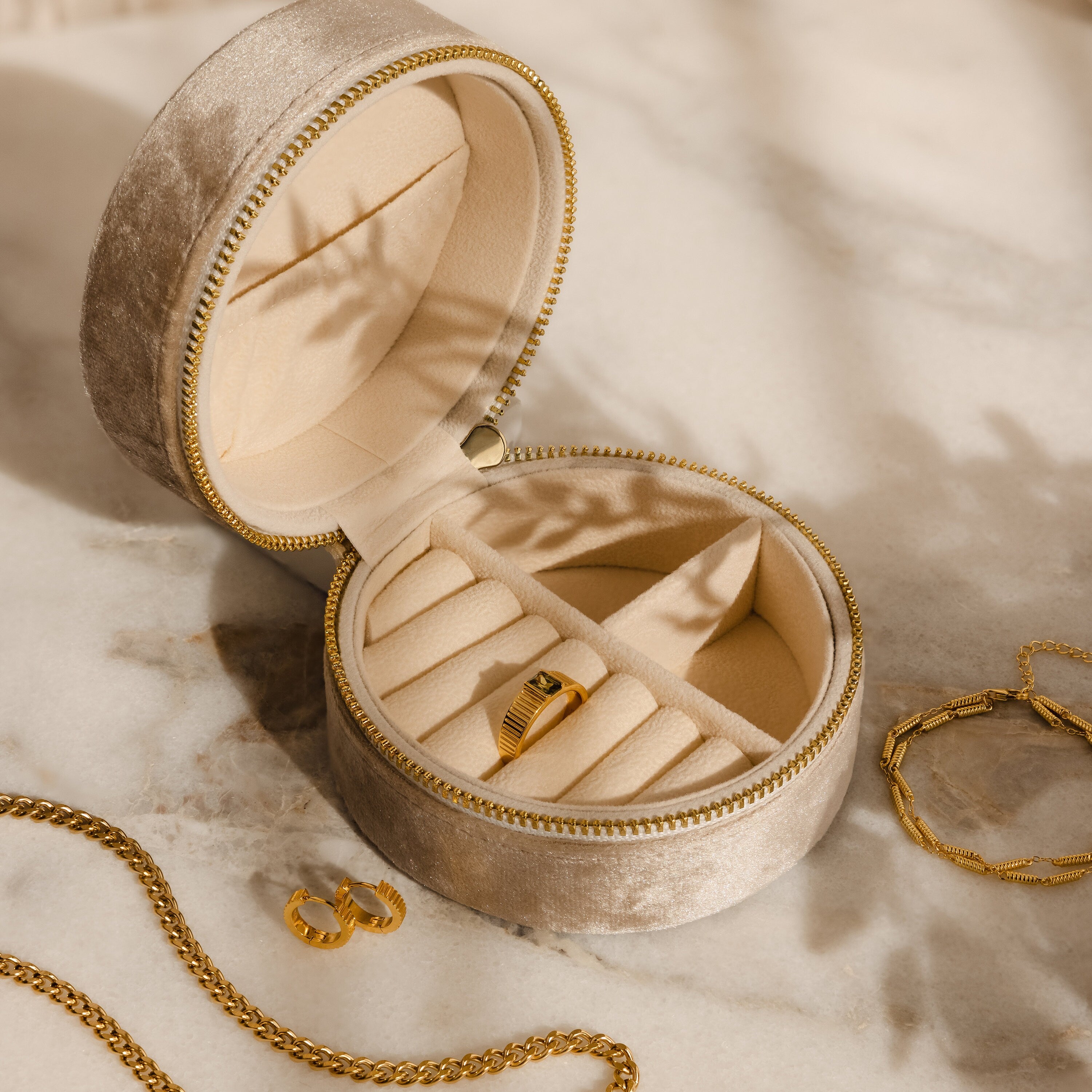 The Mystery Jewelry Box, a round beige case with handpicked gold rings and chains, rests on a marble surface bathed in warm natural light.