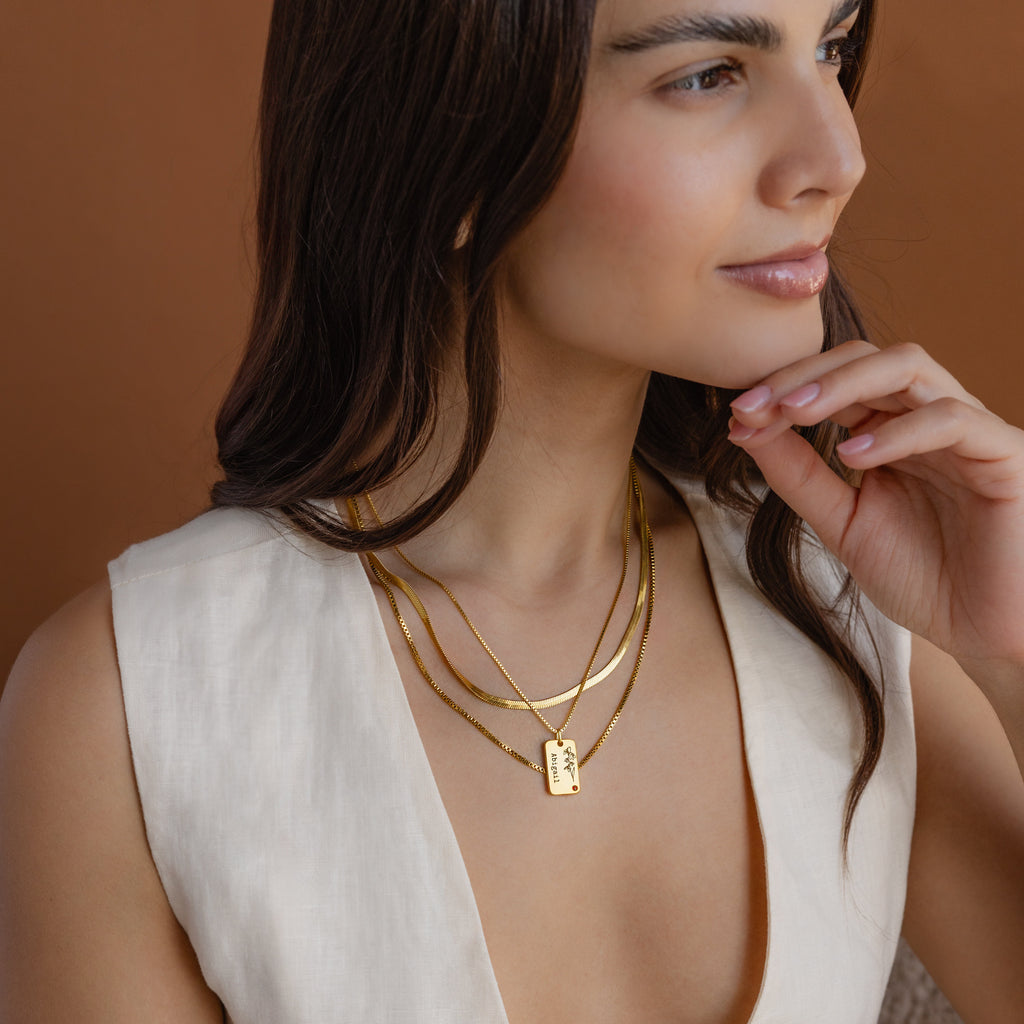 Woman wearing layered gold necklaces, including an elegant Floral Name Birthstone Necklace, and a sleeveless cream top, looking to the side against a brown background.