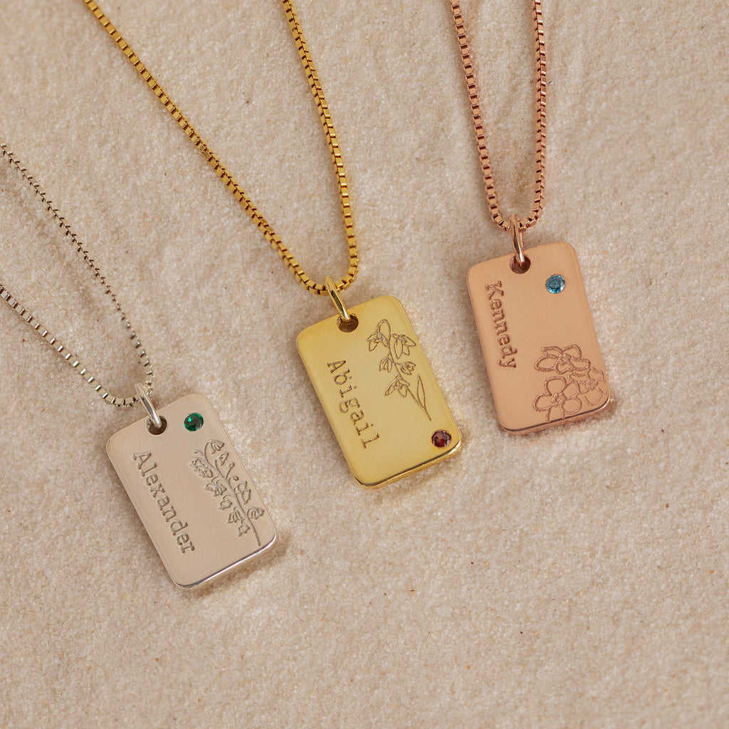 Three engraved rectangular "Floral Name Birthstone Necklaces," featuring names, flowers, and gemstones, rest on sand—a beautiful blend of personalized style.
