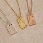 Three engraved rectangular "Floral Name Birthstone Necklaces," featuring names, flowers, and gemstones, rest on sand—a beautiful blend of personalized style.
