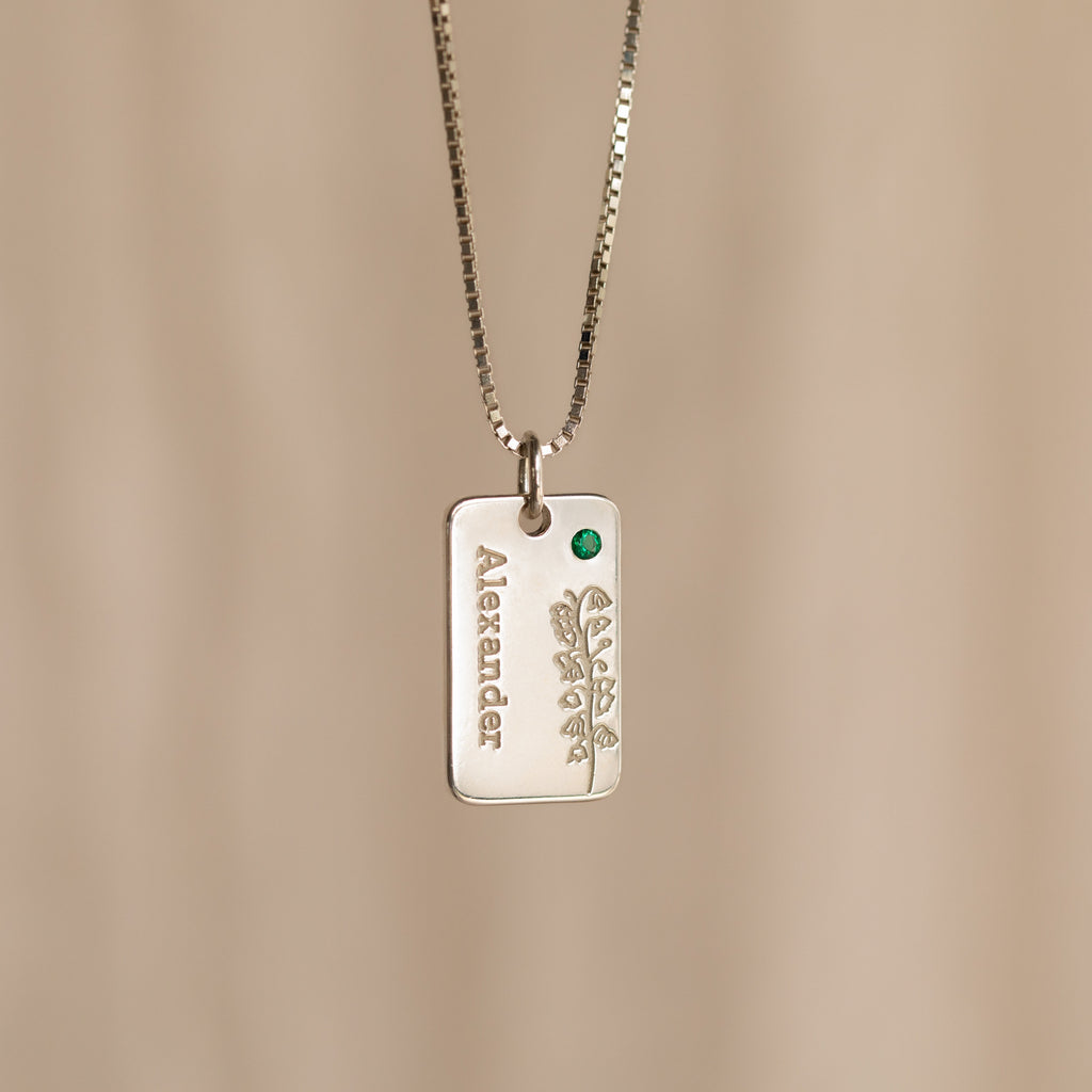 The Floral Name Birthstone Necklace features a silver rectangular pendant with a delicate leaf motif, a green gemstone, and personalized name—an elegant custom piece ideal as a birthstone necklace gift.
