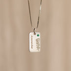The Floral Name Birthstone Necklace features a silver rectangular pendant with a delicate leaf motif, a green gemstone, and personalized name—an elegant custom piece ideal as a birthstone necklace gift.