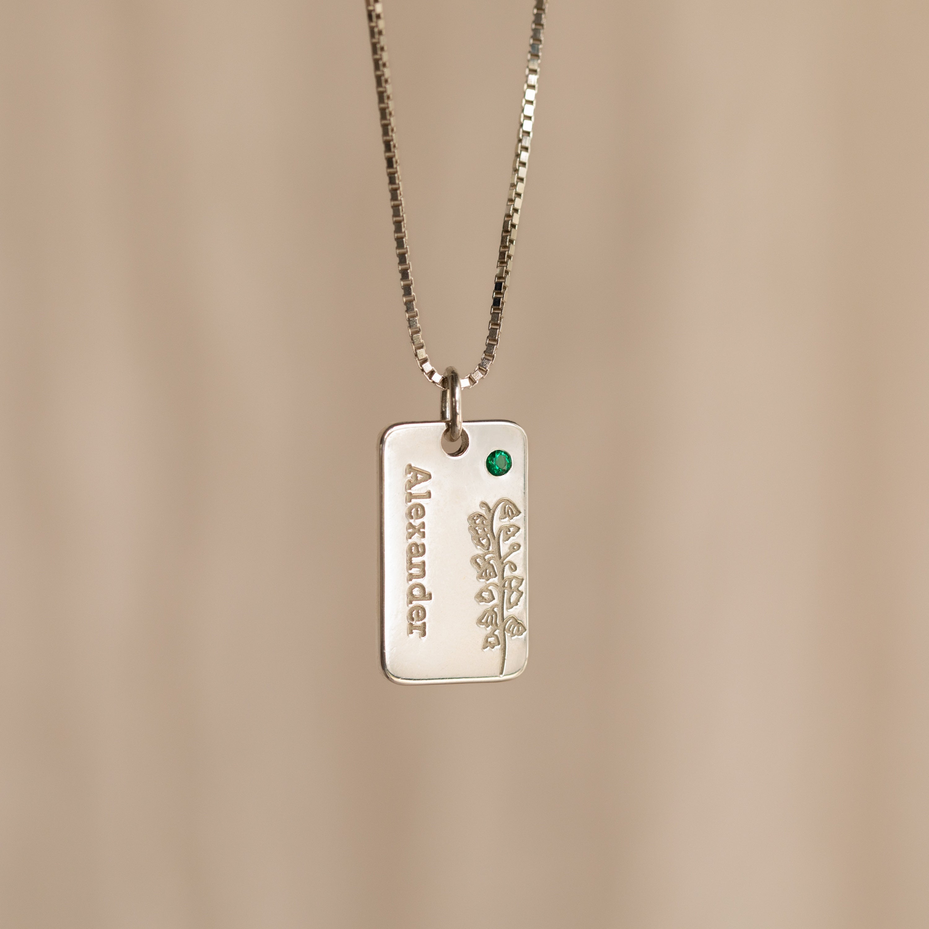 The Floral Name Birthstone Necklace features a silver rectangular pendant with a delicate leaf motif, a green gemstone, and personalized name—an elegant custom piece ideal as a birthstone necklace gift.