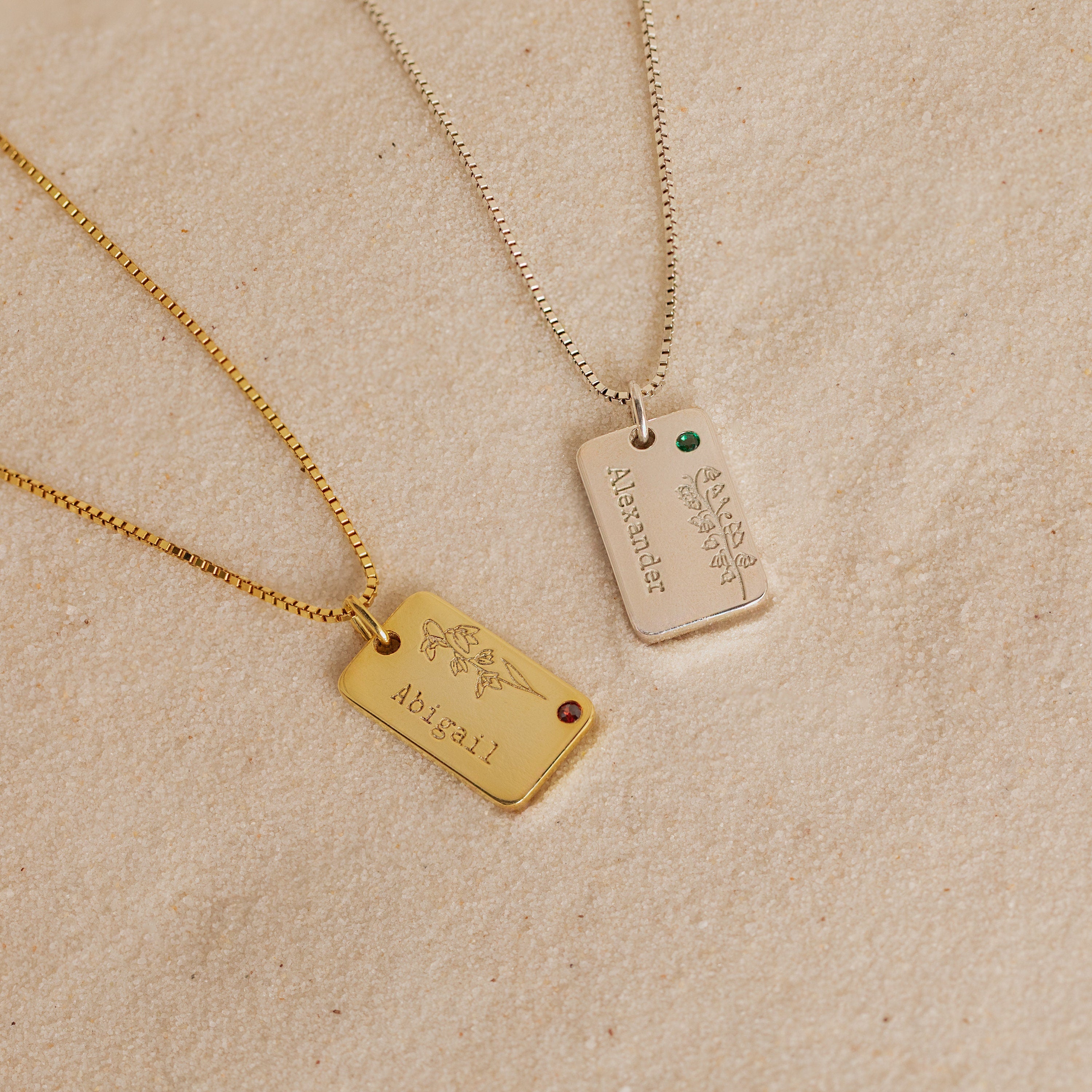 Two engraved necklaces, one gold and one silver, rest on sand—each a Floral Name Birthstone Necklace with a delicate gemstone, perfect for adding a personal touch to your style.