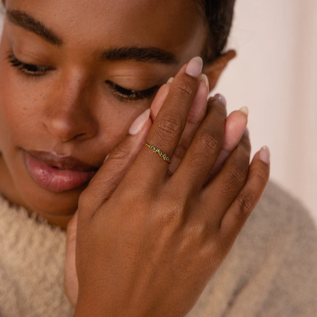 A woman with natural makeup touches her face while wearing the elegant, stackable Tiny Cluster Birthstone Ring in delicate green on her finger.