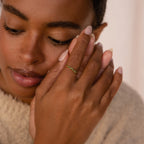 A woman with natural makeup touches her face while wearing the elegant, stackable Tiny Cluster Birthstone Ring in delicate green on her finger.