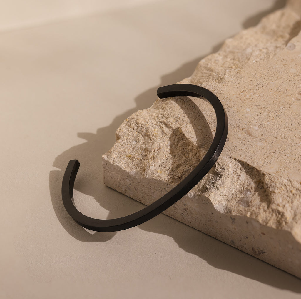 The Men's Black Cuff Bracelet is displayed on textured beige stone under soft, natural light—a stylish men’s accessory and an ideal gift for him.