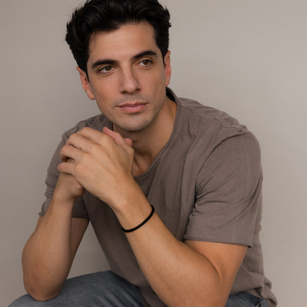 A man with dark hair in a brown t-shirt sits, hands clasped to display the Men's Black Cuff Bracelet, gazing thoughtfully to the side—an ideal gift for him.