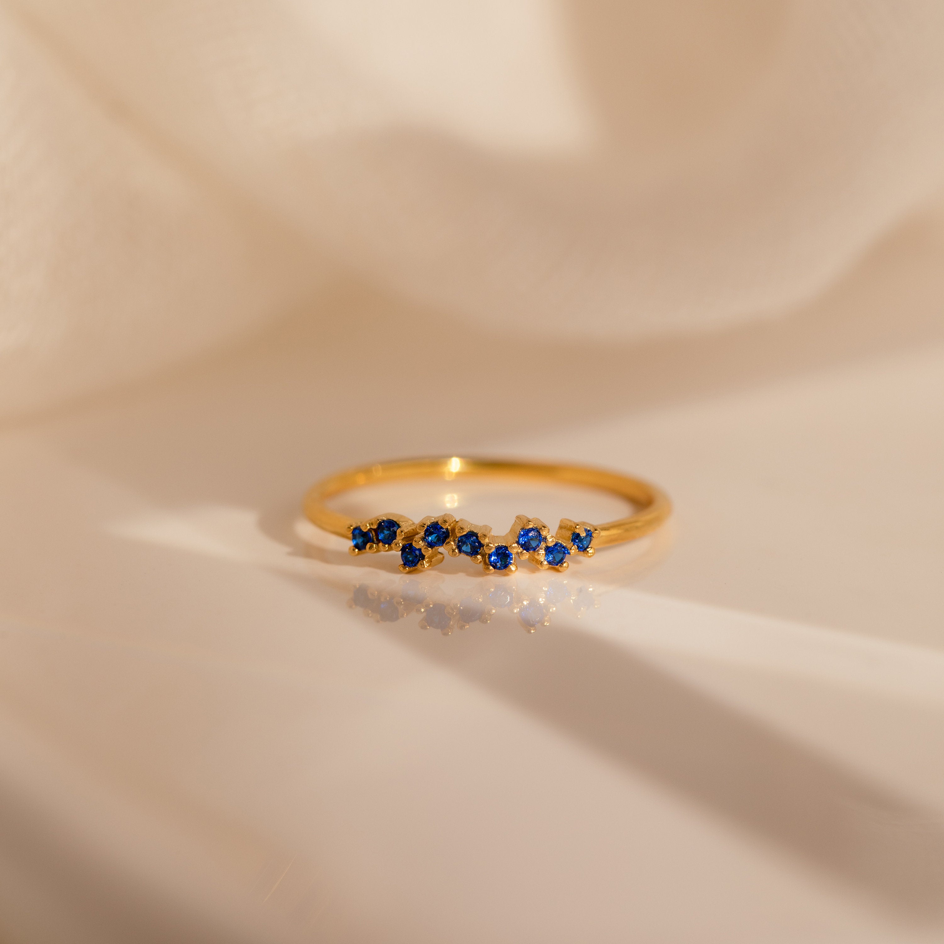The Tiny Cluster Birthstone Ring features small blue gemstones set in gold, displayed on a soft fabric backdrop—perfect as a delicate, stackable customizable ring.