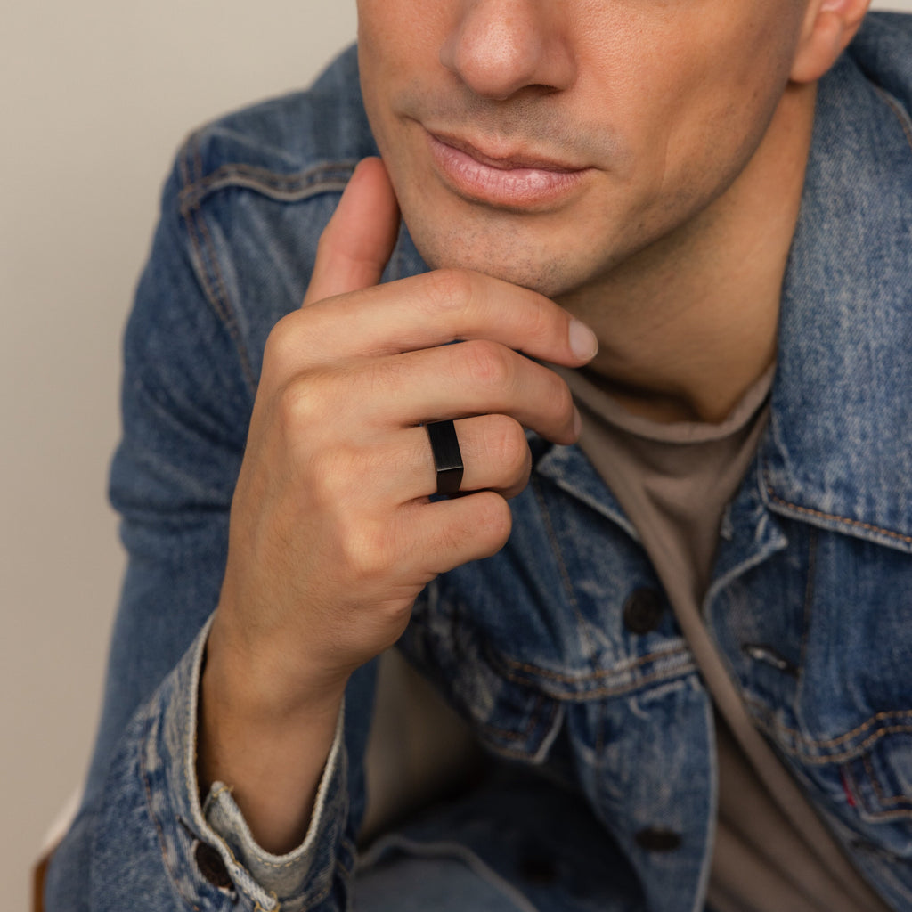 A man in a denim jacket touches his chin, showcasing the Men's Black Signet Ring on his finger.
