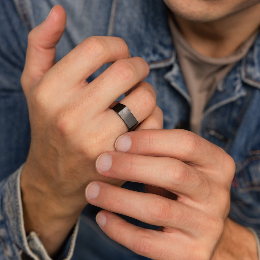 A man in a denim jacket uses both hands to adjust the Men's Black Signet Ring on his finger.