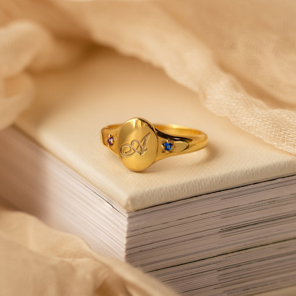The Initial Birthstone Signet Ring features a personalized initial, engraved butterfly design, and small colored stones—an elegant custom gift resting on books with beige fabric, perfect for any occasion.