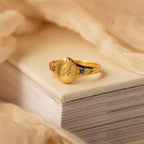 The Initial Birthstone Signet Ring features a personalized initial, engraved butterfly design, and small colored stones—an elegant custom gift resting on books with beige fabric, perfect for any occasion.