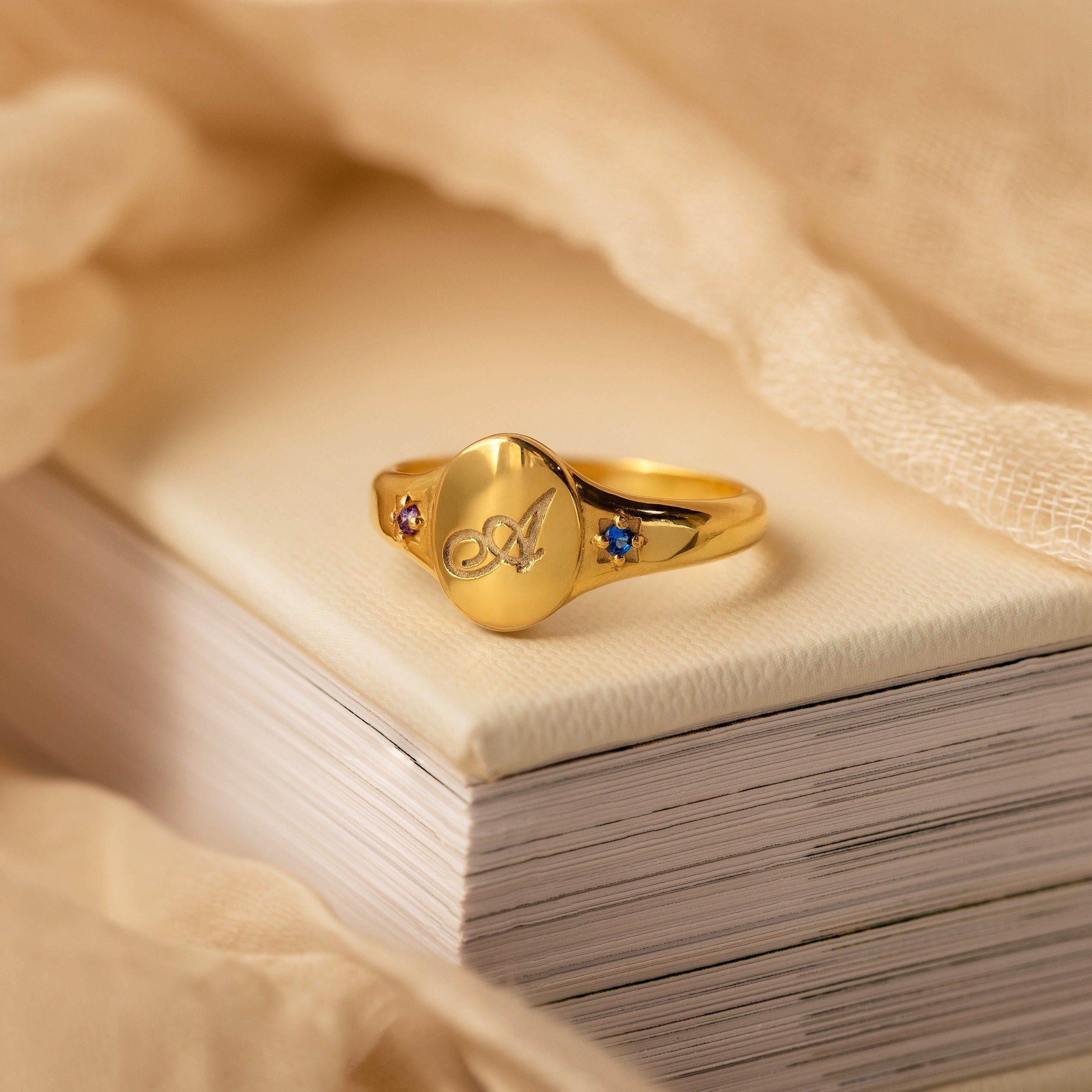 The Initial Birthstone Signet Ring features a personalized initial, engraved butterfly design, and small colored stones—an elegant custom gift resting on books with beige fabric, perfect for any occasion.