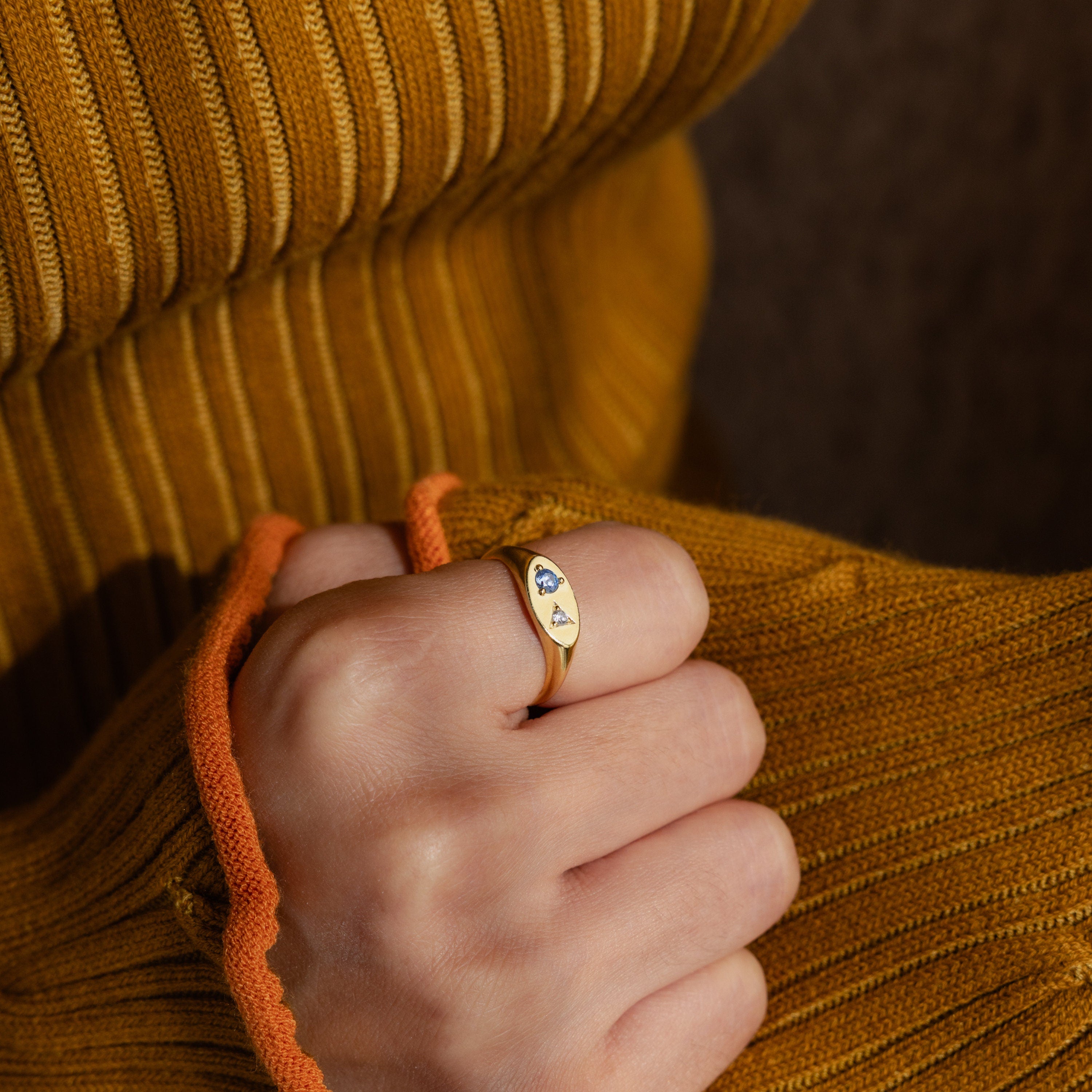 A hand in a mustard sweater showcases the Mixed Birthstone Signet Ring, a gold statement piece featuring delicate blue and white stones.