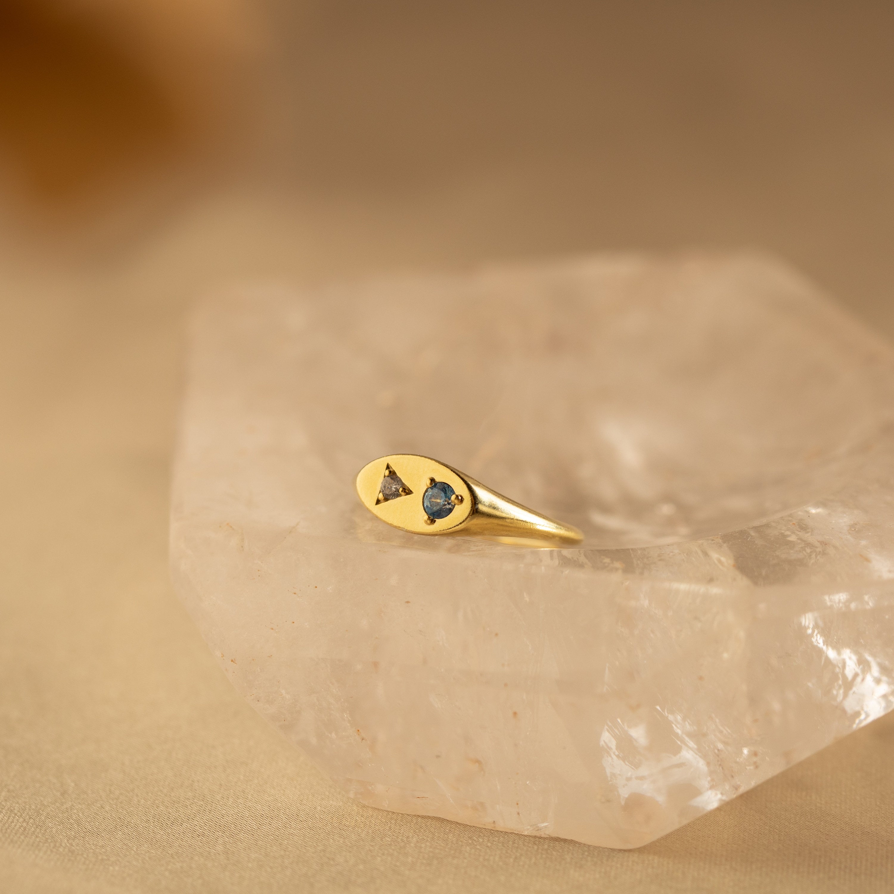 The Mixed Birthstone Signet Ring, featuring two small gemstones set in gold, is displayed on a clear quartz crystal with a beige background.