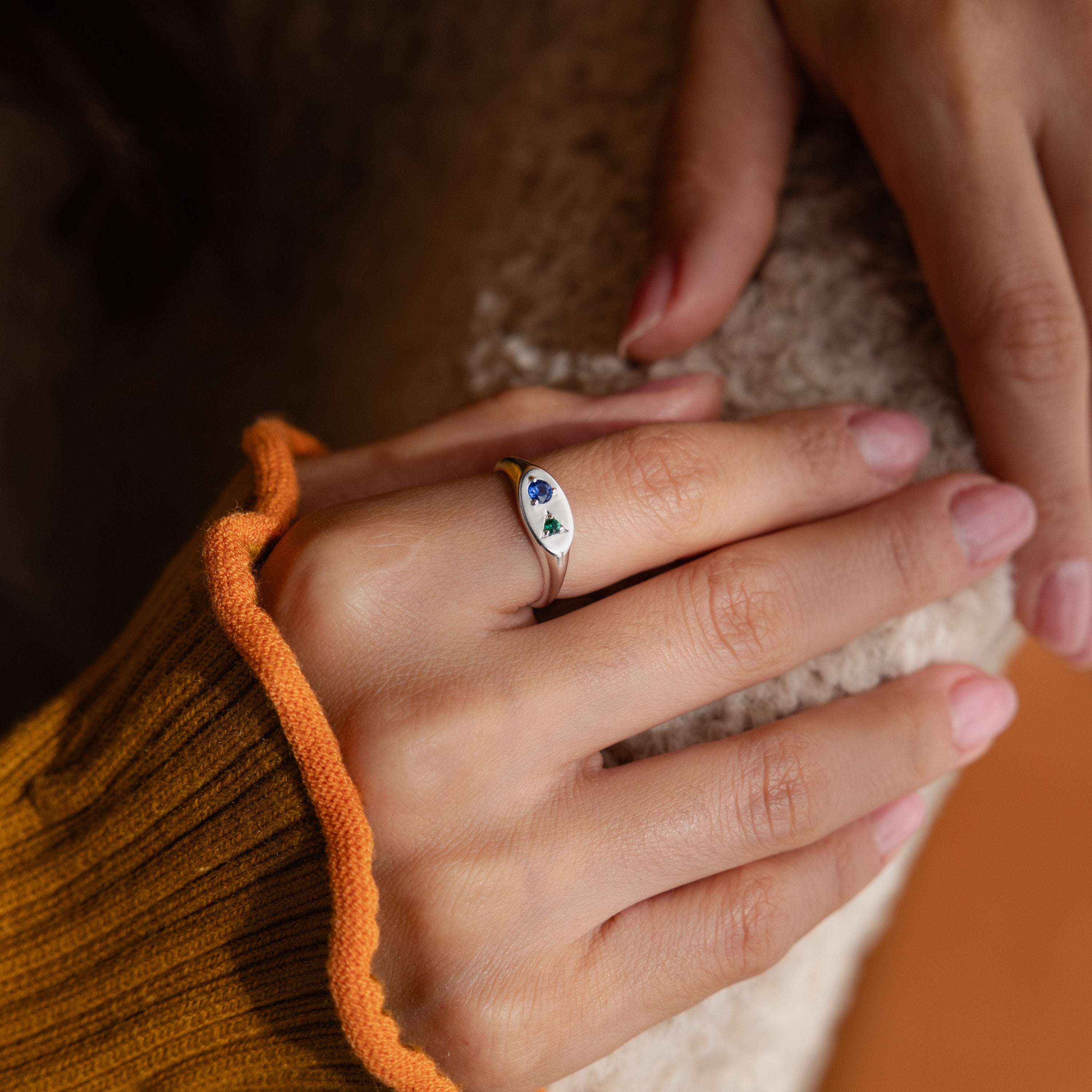 A hand with light pink nails wears the Mixed Birthstone Signet Ring, featuring blue and green accents, resting on a fuzzy surface for a unique statement look.