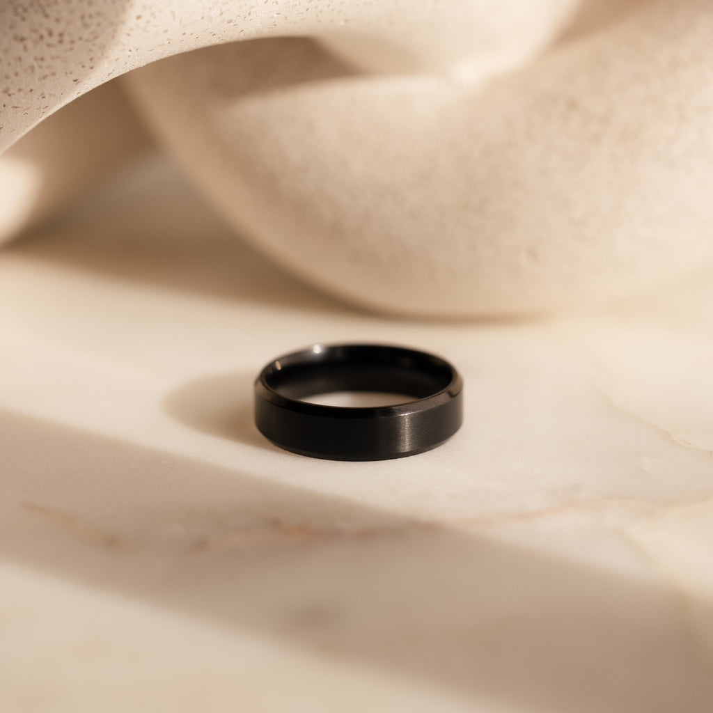The Men's Thick Black Ring sits on a smooth, light marble surface with abstract shapes in the background, showcasing its bold and modern elegance.