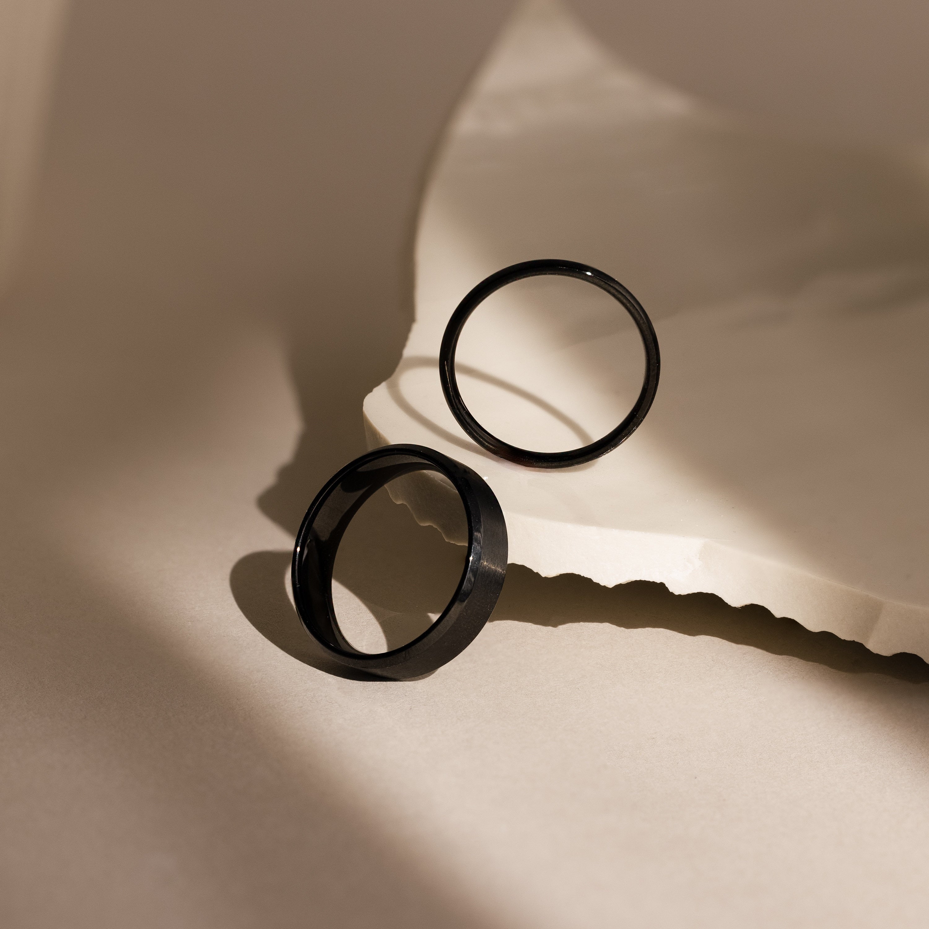 The Men's Thick Black Rings, perfect as men's rings or a modern wedding band, are displayed on a textured beige surface with soft shadows and abstract lighting.