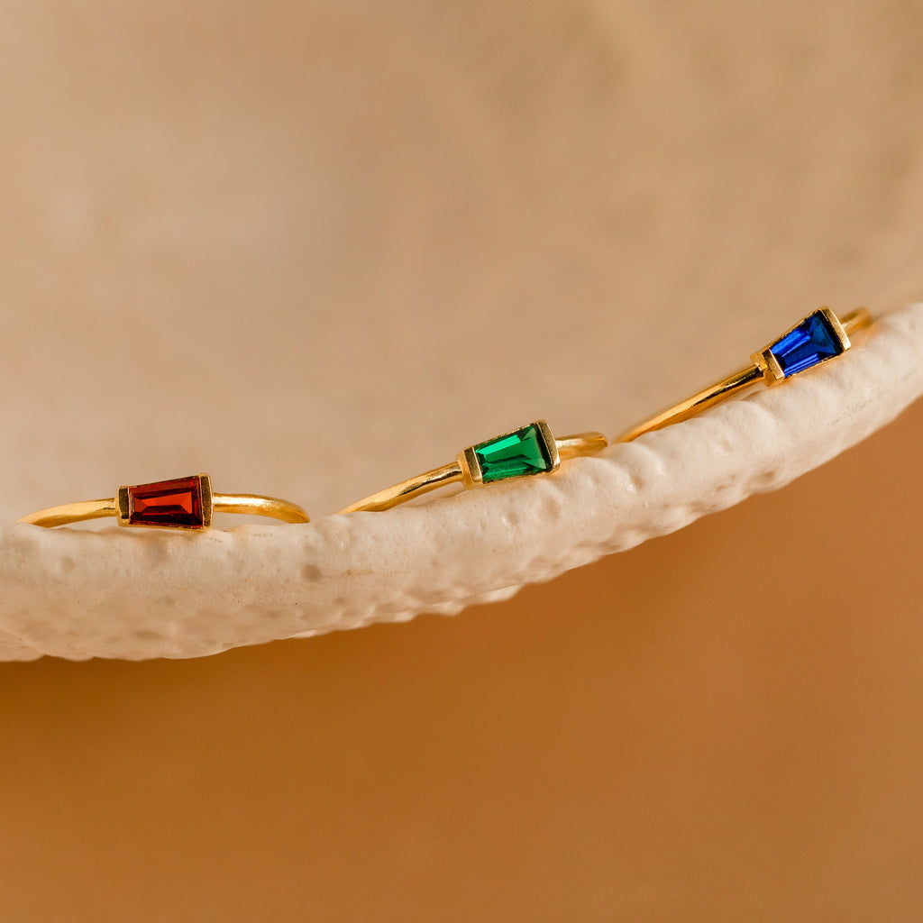 Three gold rings, each featuring a red, green, or blue gemstone, rest on a textured beige dish—each one an Irregular Baguette Birthstone Ring, ideal for creating your own unique and customized ring stack.