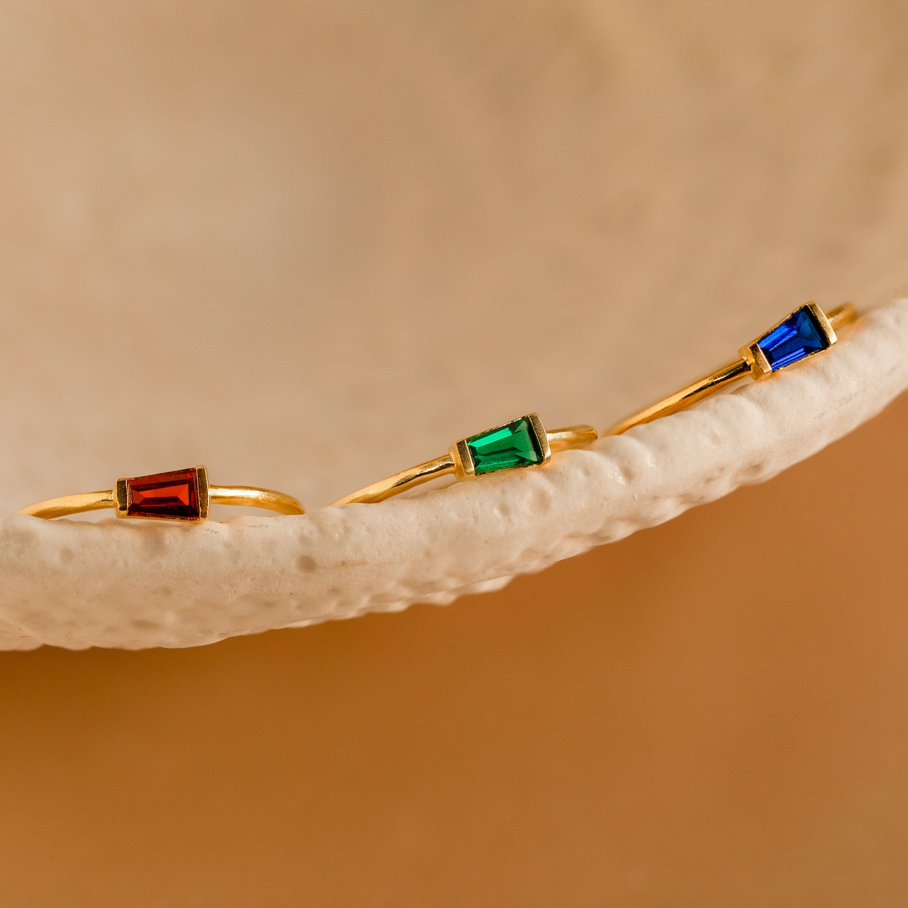 Three gold rings, each featuring a red, green, or blue gemstone, rest on a textured beige dish—each one an Irregular Baguette Birthstone Ring, ideal for creating your own unique and customized ring stack.