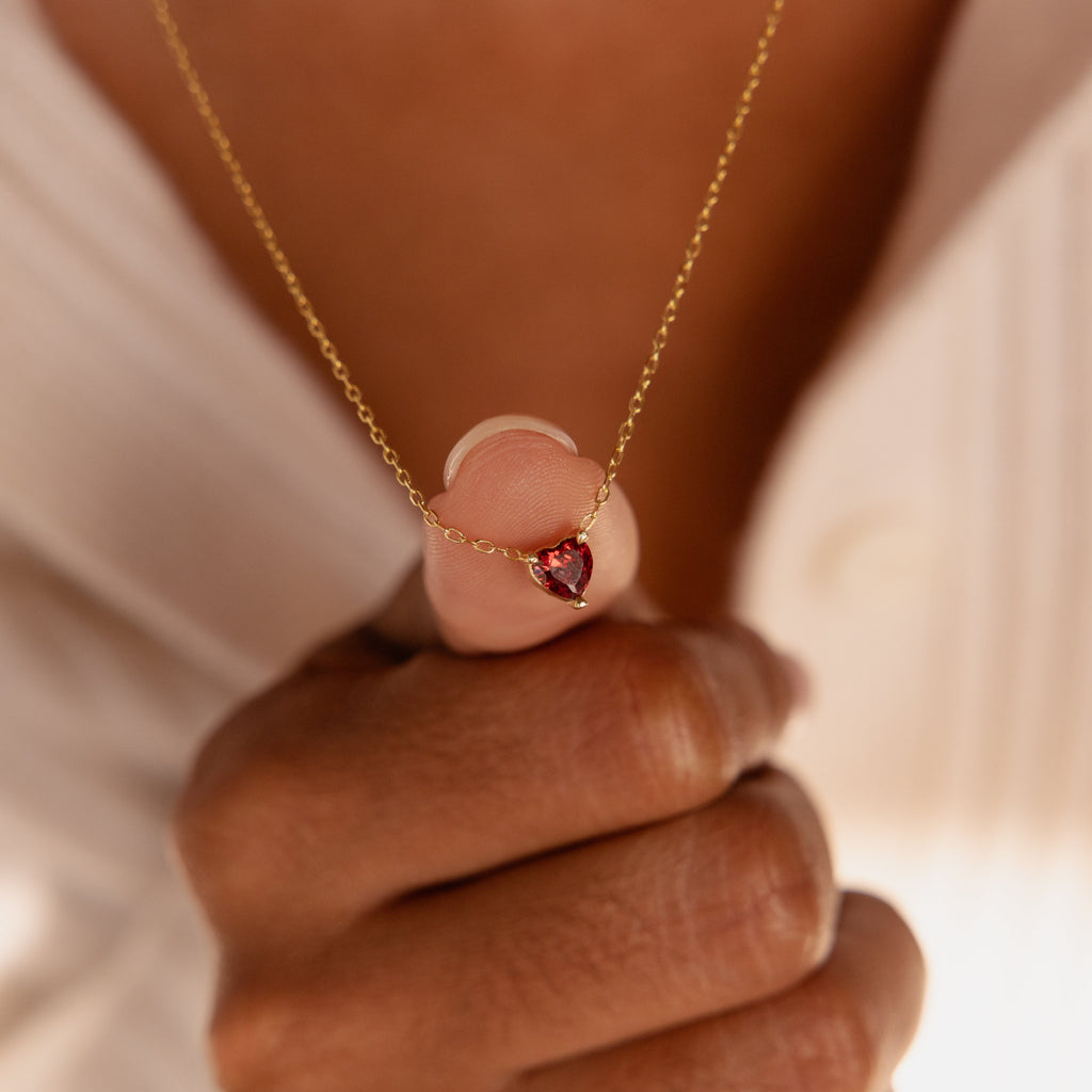 A hand holds the Heart Birthstone Necklace featuring a small gold red heart-shaped pendant, shown over a cream-colored top—an ideal personalized jewelry piece.