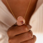 A hand holds the Heart Birthstone Necklace featuring a small gold red heart-shaped pendant, shown over a cream-colored top—an ideal personalized jewelry piece.